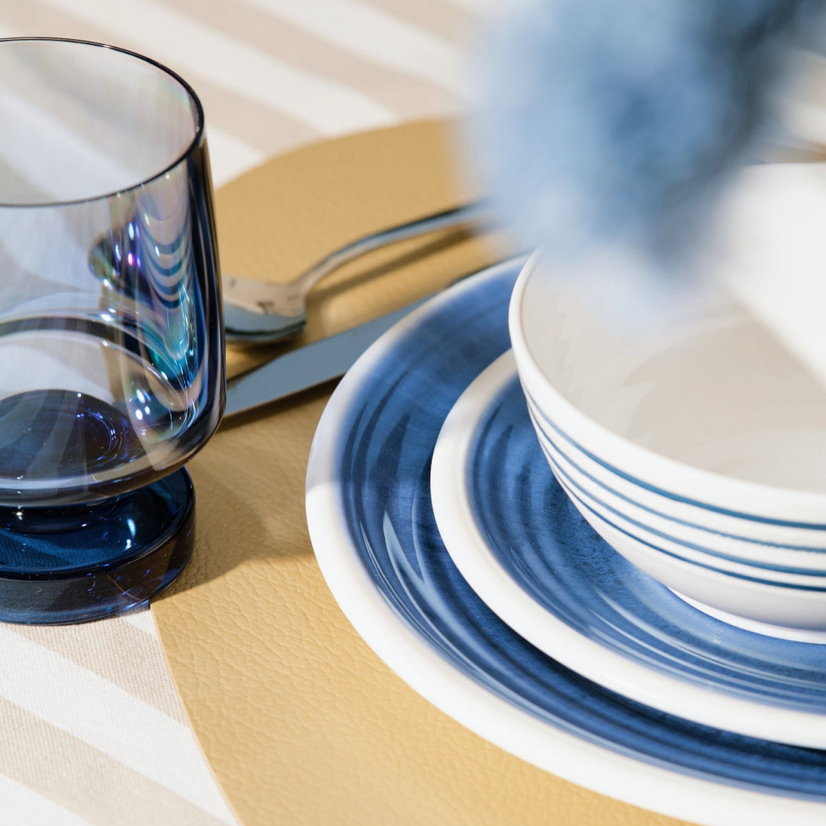 Flamefield sfeerbeeld van onbreekbaar servies Azure blauw met wit AZ0112 op een rieten placemat met een witte mok en bestek  met blauwe wijnglas Heavy Base ingezoomd