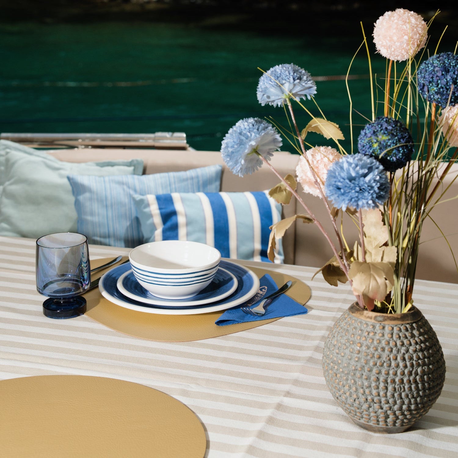 Flamefield sfeerbeeld van onbreekbaar servies Azure blauw met wit AZ0112 op een rieten placemate met een witte mok en bestek  met blauwe wijnglas Heavy en een vaas met bloemen op een boot