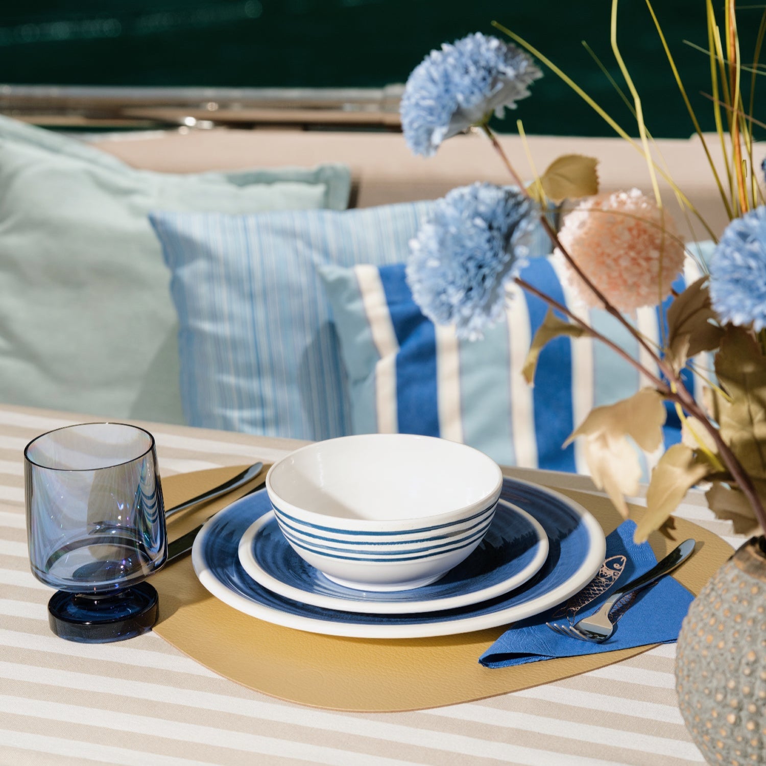 Flamefield sfeerbeeld van onbreekbaar servies Azure blauw met wit AZ0112 op een rieten placemate met een witte mok en bestek  met blauwe wijnglas Heavy en een vaas met bloemen op een boot ingezoomd