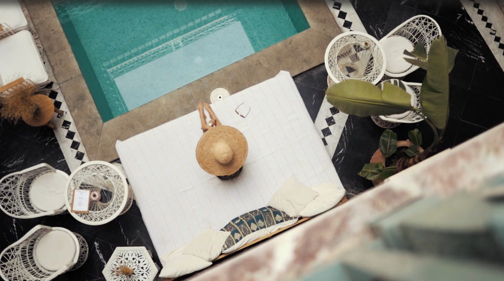 Studio_Maritime_live_the_sea_love_the_style_hompeage beeld van zwembad van bovenaf met vrouw met rieten houd en wit kleed en witte rotan stoelen en tafel
