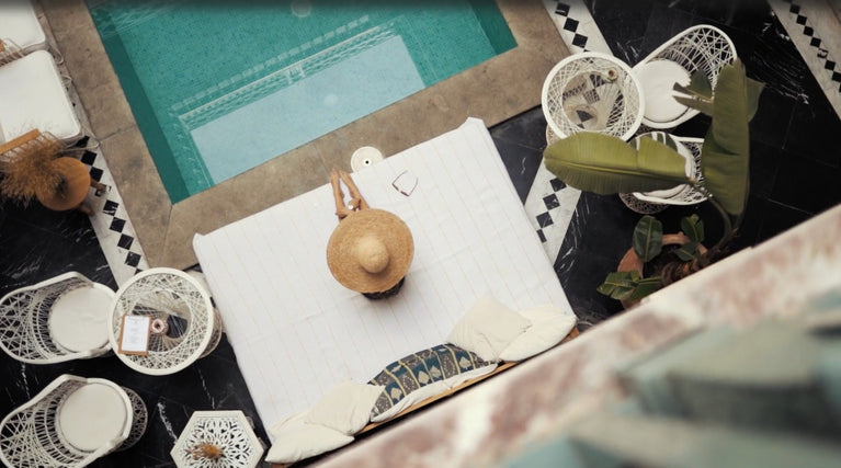 Studio_Maritime_live_the_sea_love_the_style_hompeage beeld van zwembad van bovenaf met vrouw met rieten houd en wit kleed en witte rotan stoelen en tafel