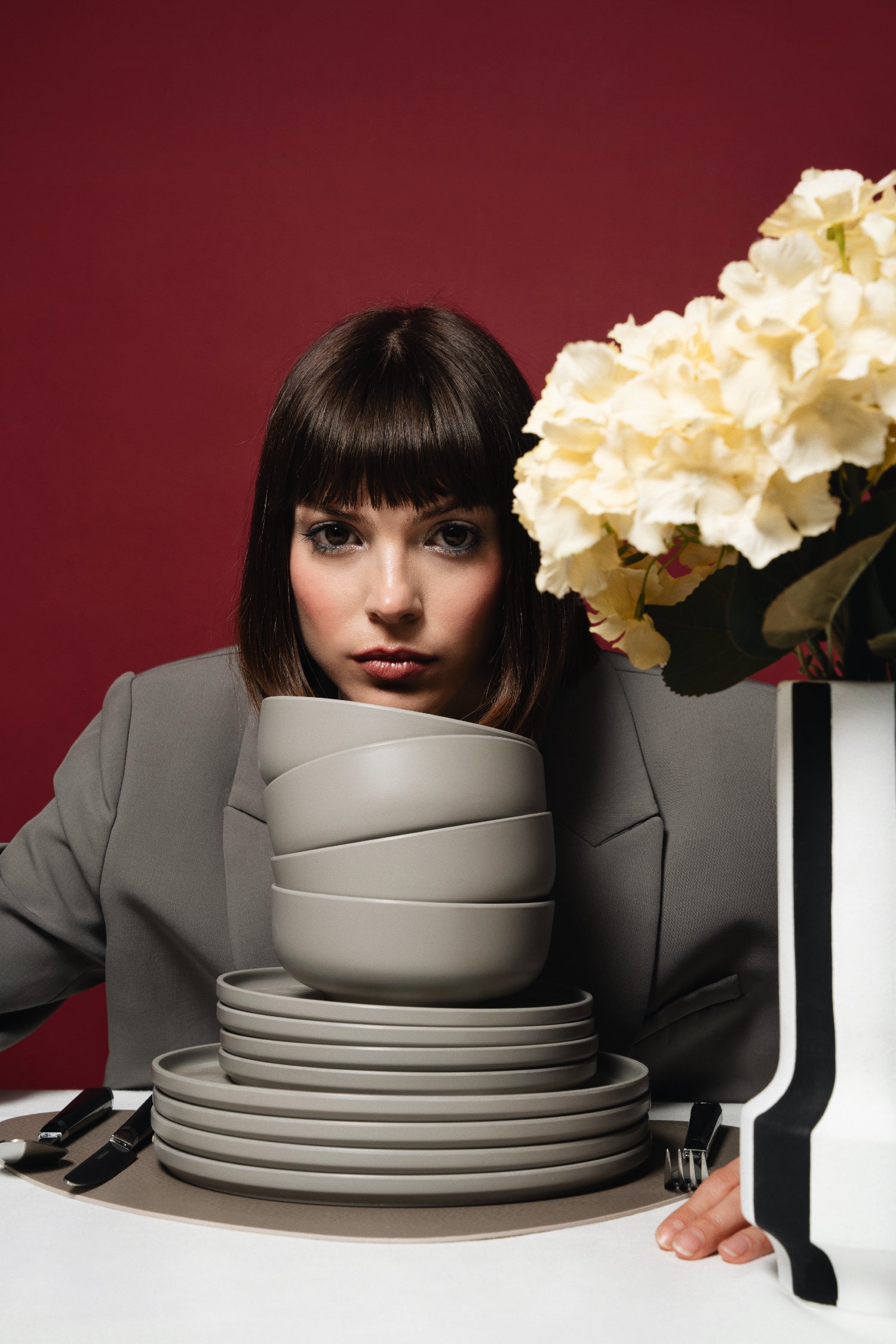 Vrouwelijk model met Flamefield serviesset Pebble Sky – 12-delig, mat grijs melamine set en een vaas met bloemen tegen bordeaux rode achtergrond